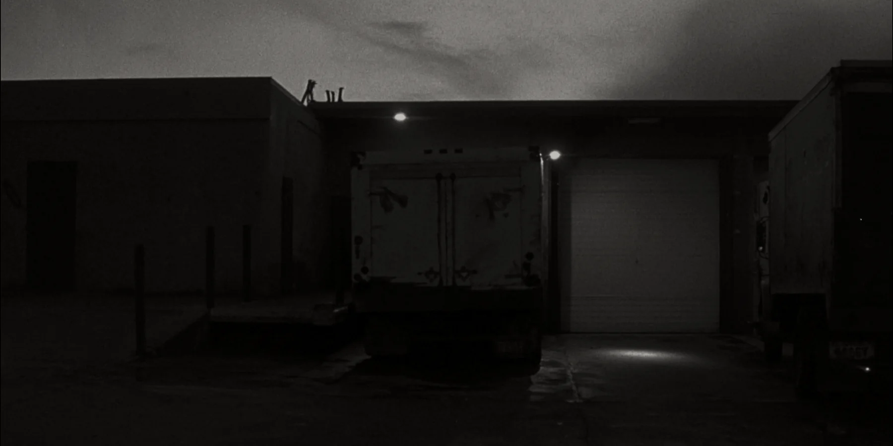 A plain box truck backed square into a loading dock before sunrise, roll door up and the dock light throwing a hard pool onto the wet apron.