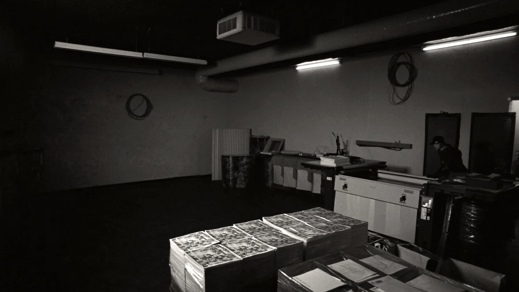 A sheetfed press running at night on the floor of a small print and mail plant, pallets of uncut sheets stacked in the foreground.