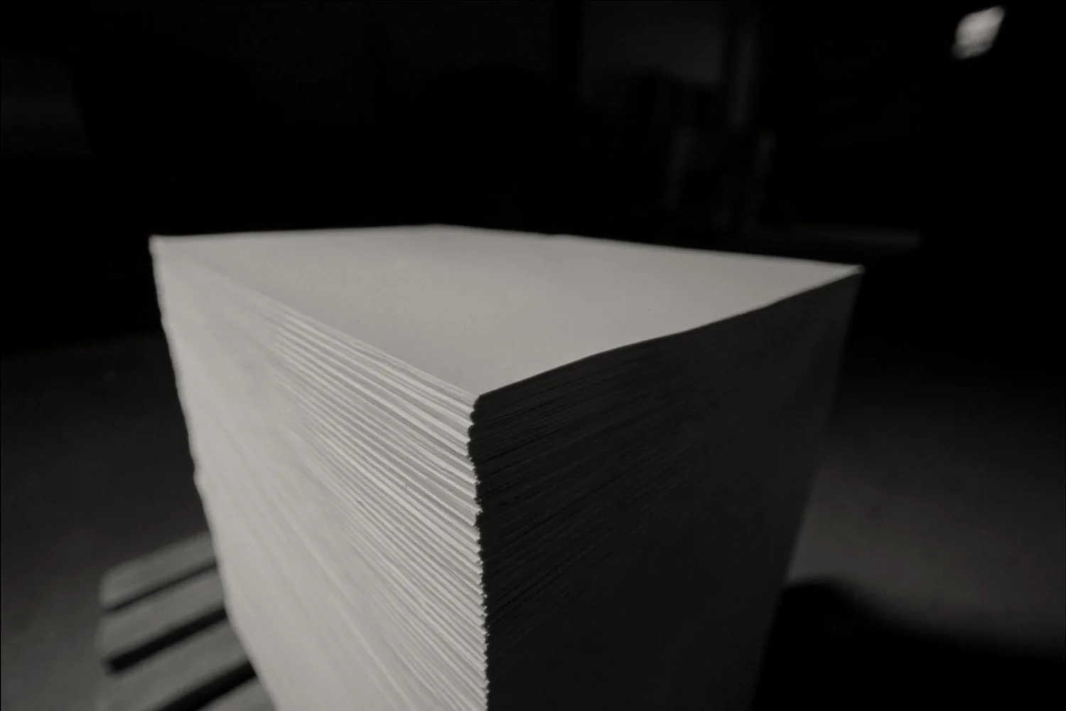 A tall stack of freshly guillotined blank postcard stock on a wooden skid, the cut edges catching a single raking light in a dark plant.