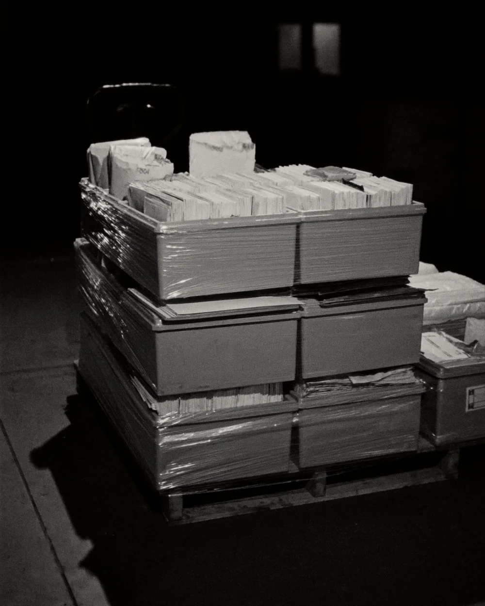 A pallet stacked shoulder high with grey plastic postal letter trays packed with upright mail, half wrapped, on a dim warehouse floor.